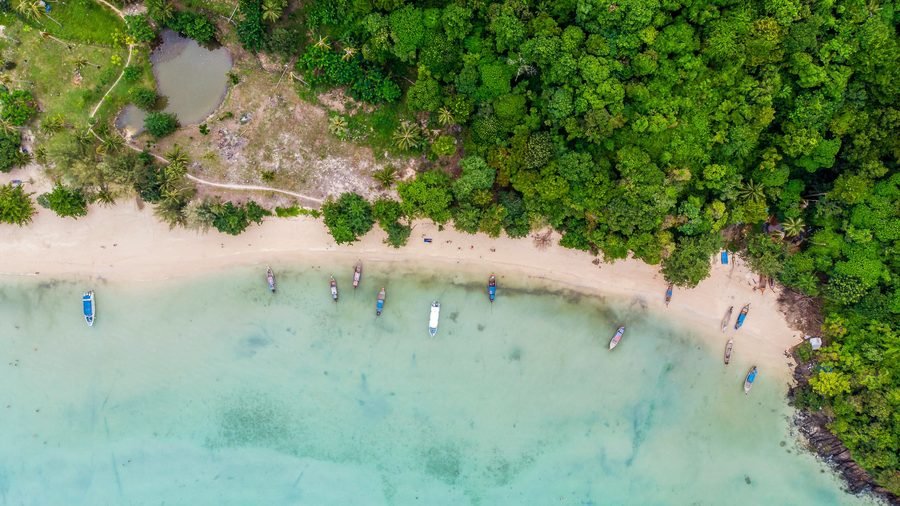 A sunlit cove from above, longtail boats moored in clear turquoise water below a forested shore