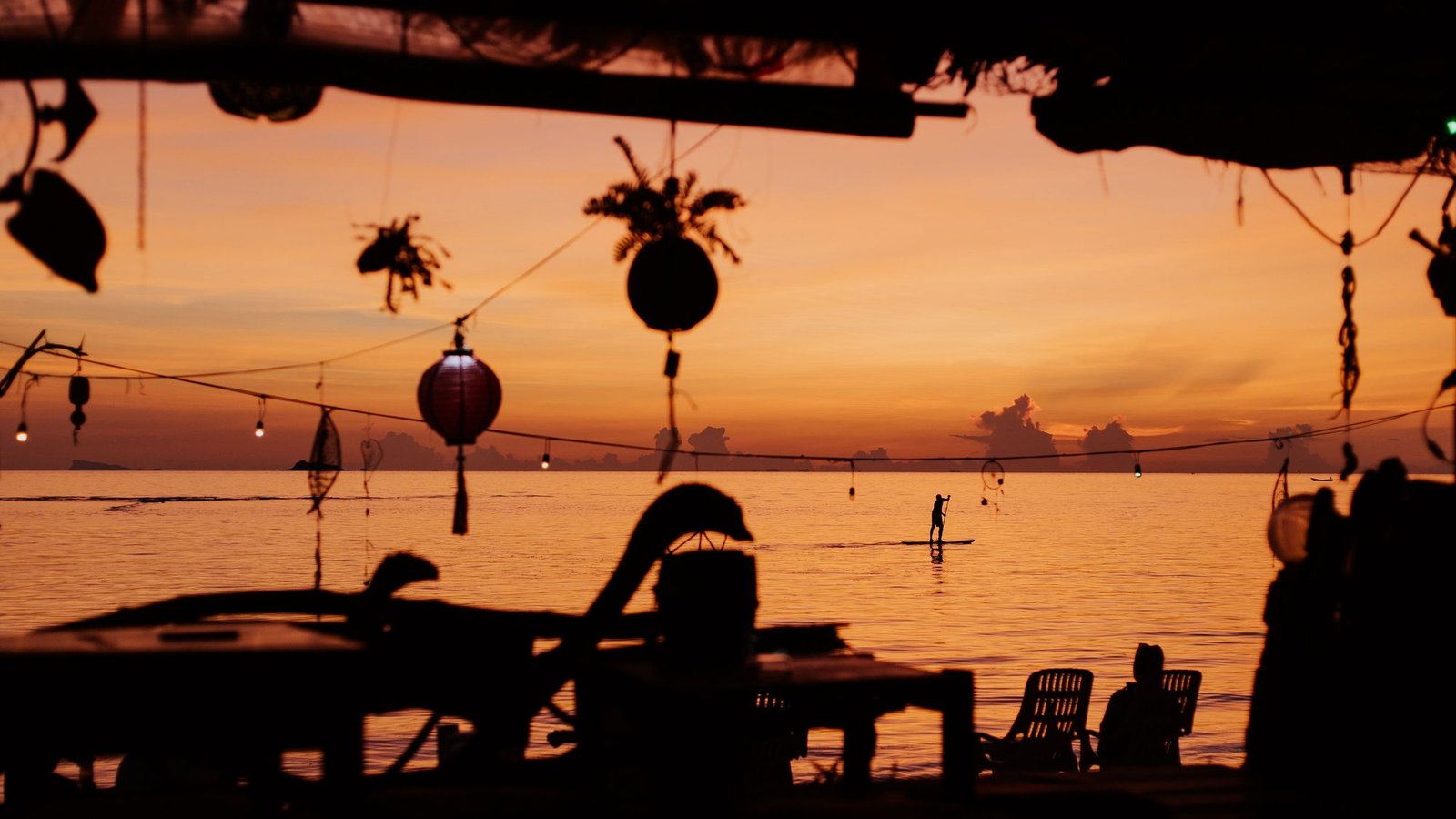 Des lanternes et des guirlandes lumineuses dans un bar de plage, les tables en contre-jour devant une mer orange après le coucher du soleil