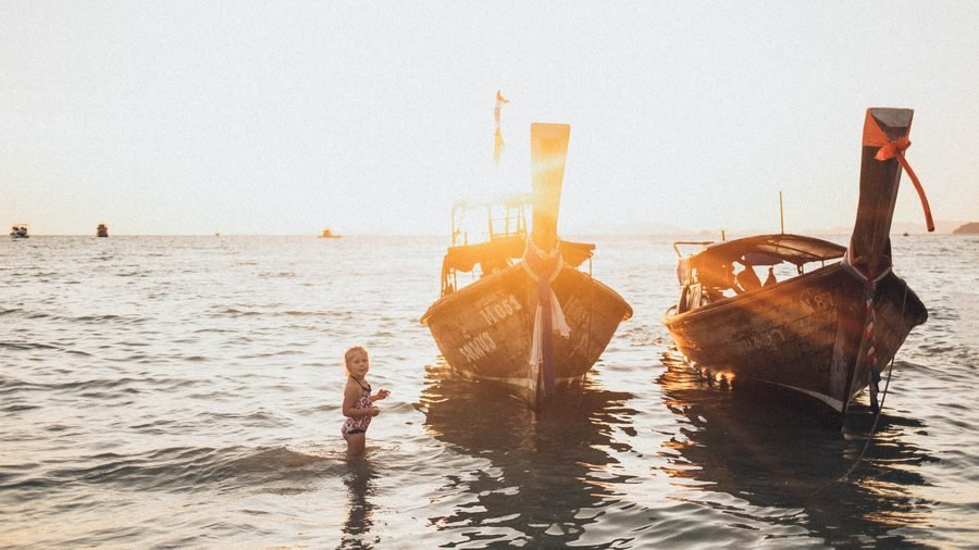 A small child paddling in the shallows beside two longtail boats at sunset