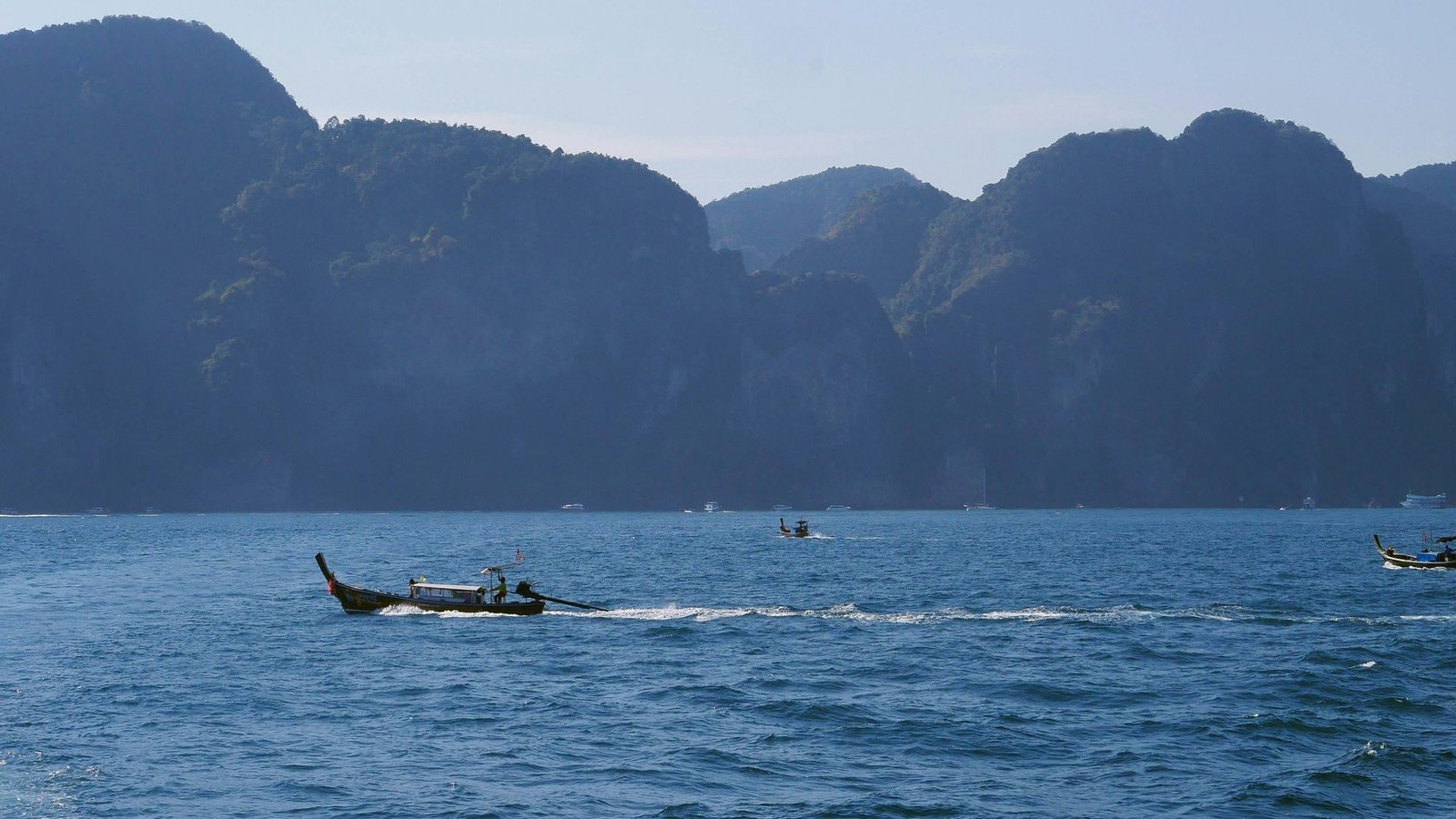 Longtail boats crossing open sea in front of tall forested limestone cliffs
