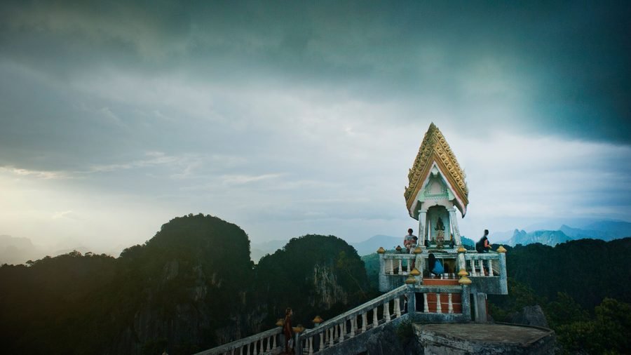 A small white and gold shrine on a hilltop terrace, surrounded by forested limestone peaks under a stormy sky