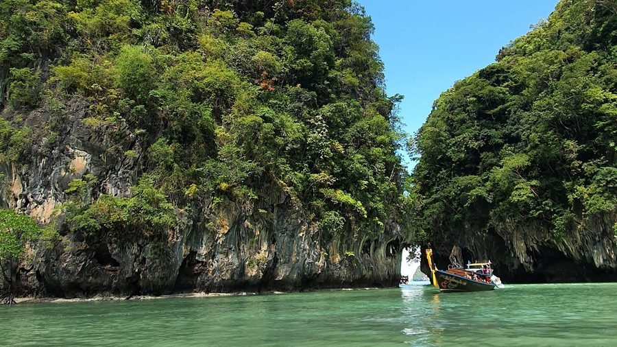 A longtail boat at the narrow entrance to a lagoon between two forested limestone cliffs