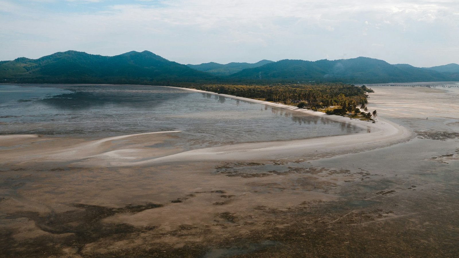Aerial view at low tide of a long white sand spit lined with palms, forested hills of Koh Yao Yai behind