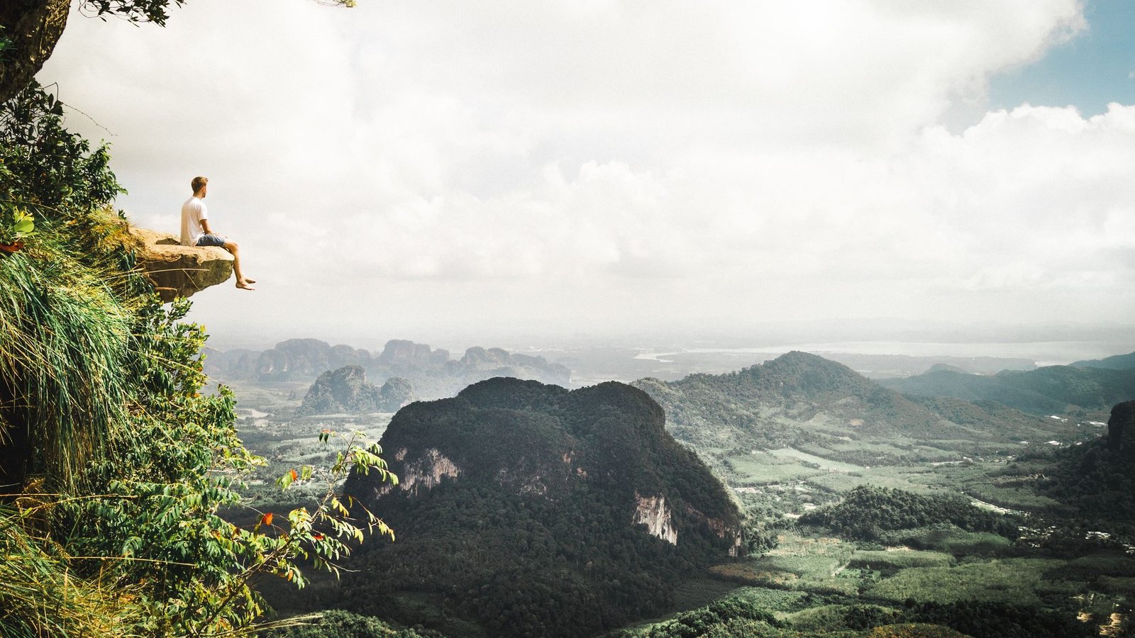 A hiker sitting on a rock ledge above a valley of limestone peaks in the haze