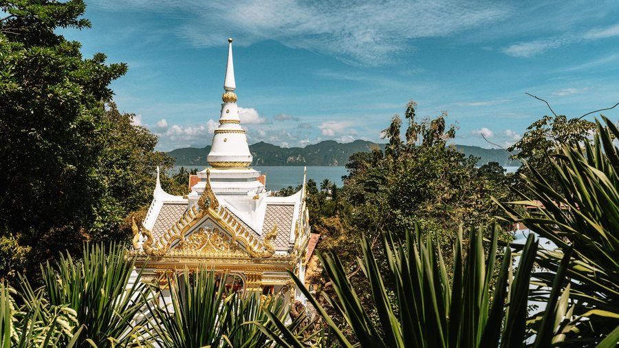 White and gold Buddhist shrine among palms, a bay and limestone hills behind, in Krabi province