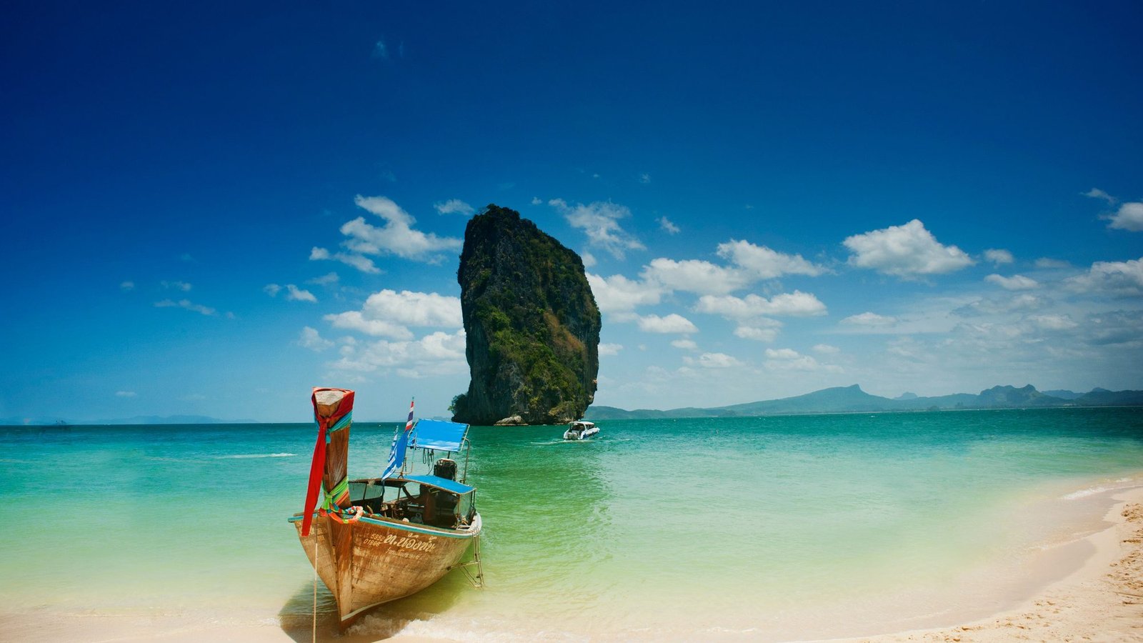 A longtail boat on a white-sand beach, with a limestone stack rising offshore