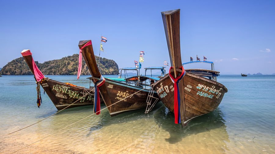 Three wooden longtail boats moored in clear shallow water, with a wooded island behind