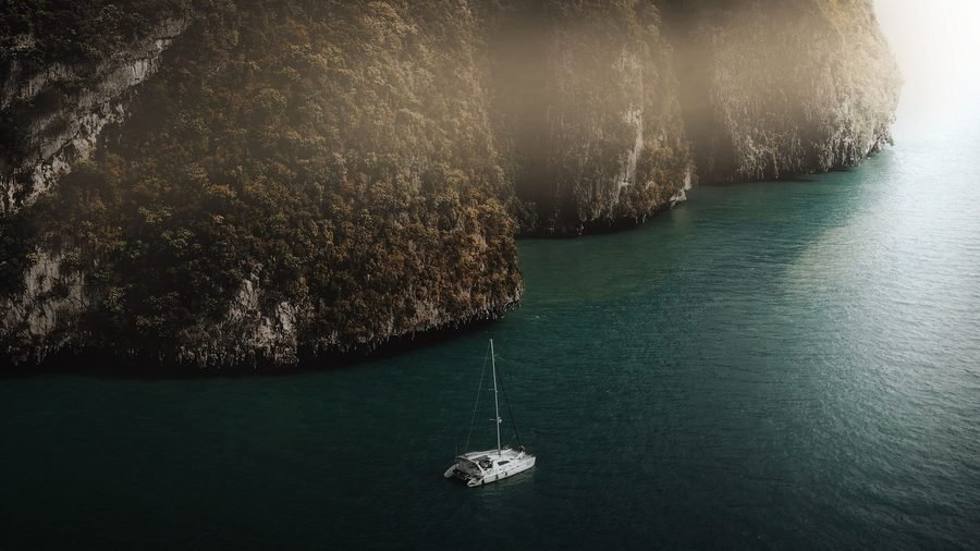 Forested limestone cliffs in a golden haze above dark green water, a catamaran anchored below, seen from the air